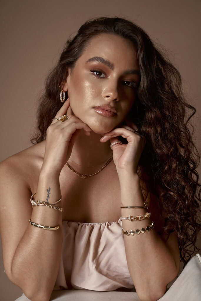 Abril Escalante a model with a radiant complexion showcases an array of gold jewelry including hoop earrings necklaces and bracelets against a muted brown backdrop.